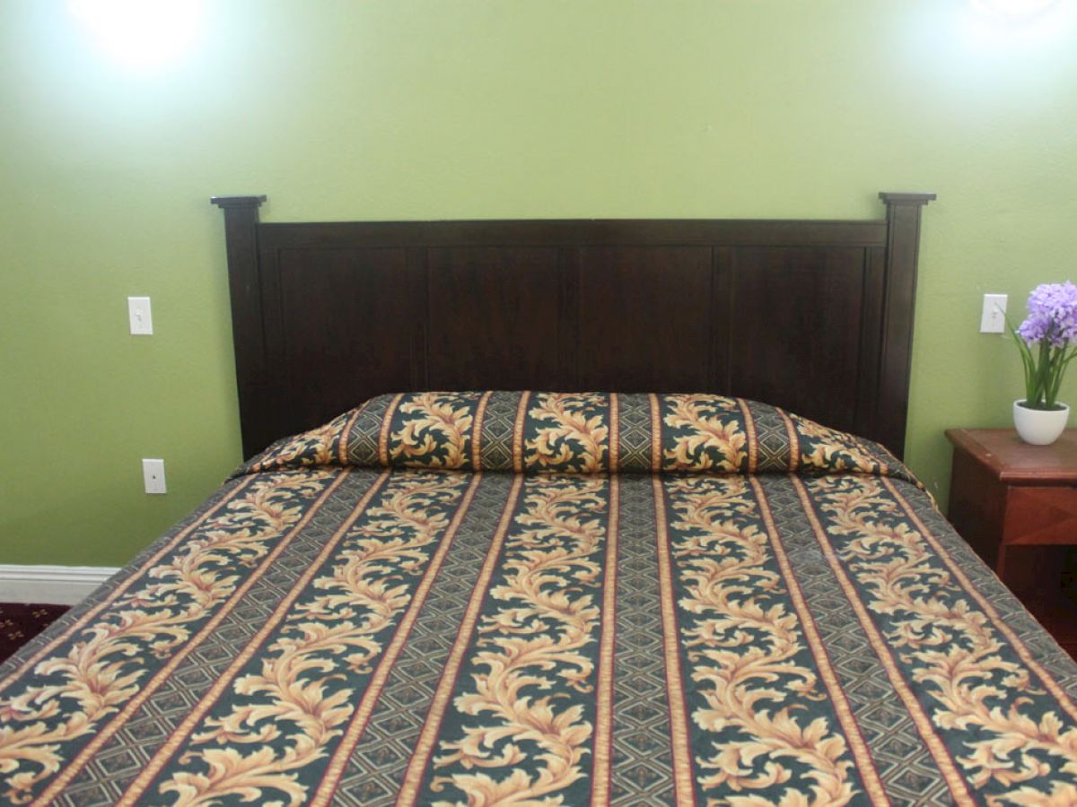 The image shows a bed with a patterned bedspread against a green wall, flanked by two nightstands, one holding a potted plant with purple flowers.