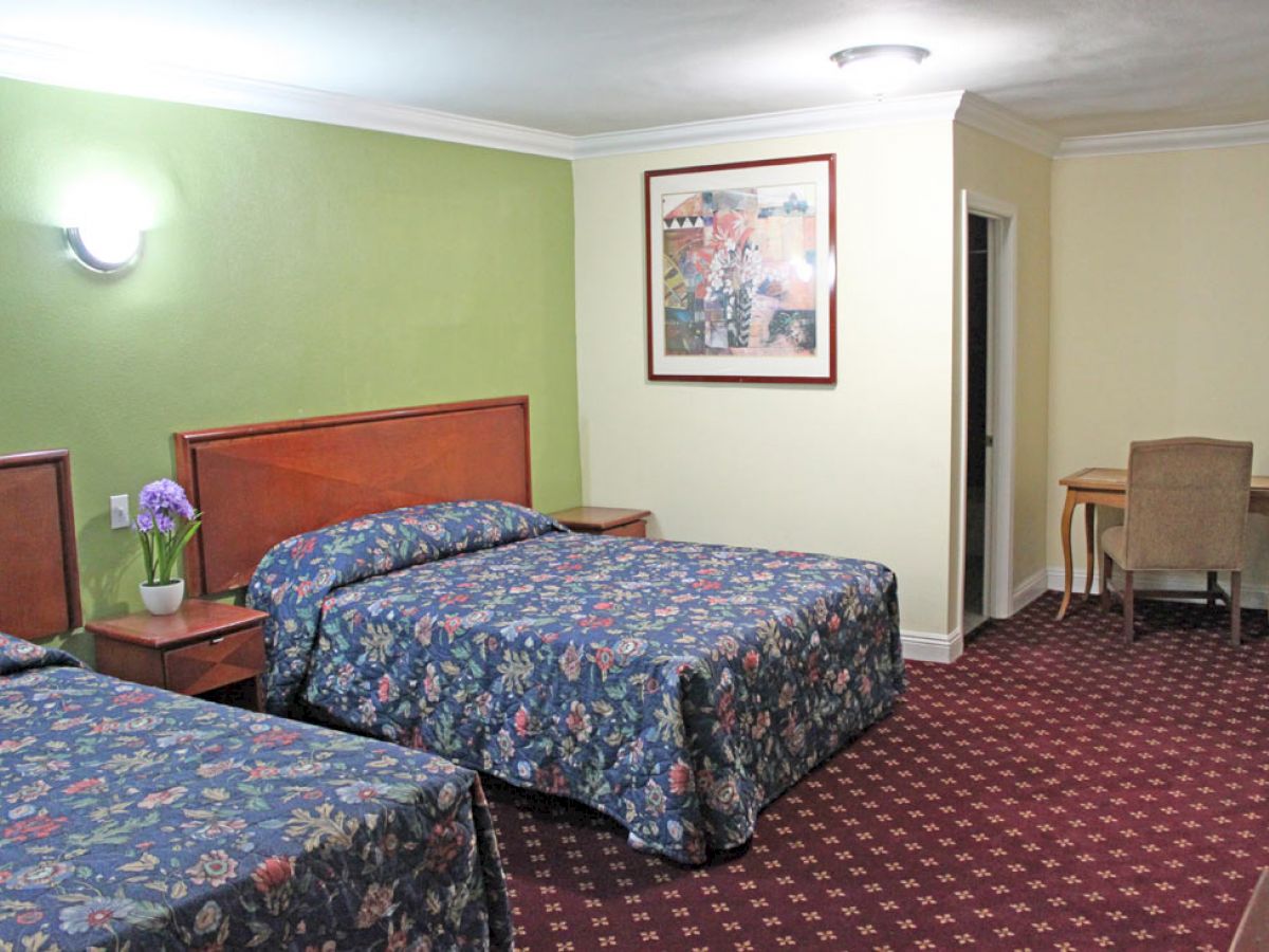 The image shows a hotel room with two beds, floral bedspreads, green and beige walls, a painting, wall lamp, nightstands, a desk, and a chair.