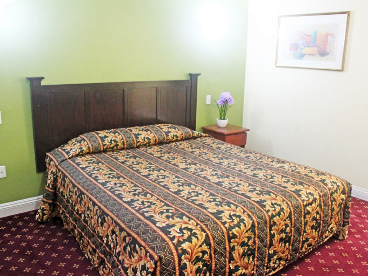 A bedroom with a double bed covered with a patterned bedspread, two side tables, a flower vase, a painting on the wall, and green and white walls.