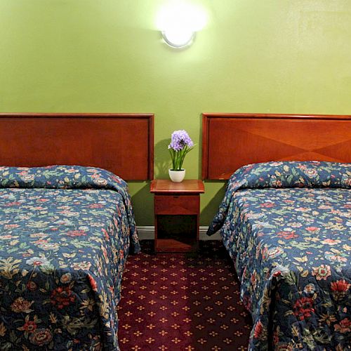 This image shows a hotel room with two beds, floral bedspreads, a nightstand in between with a vase of flowers, and green walls with wall lamps.