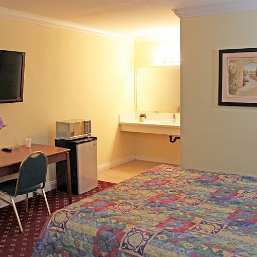 A hotel room with a bed, desk and chair, wall-mounted TV, artwork, mini-fridge, microwave, and a sink area visible.