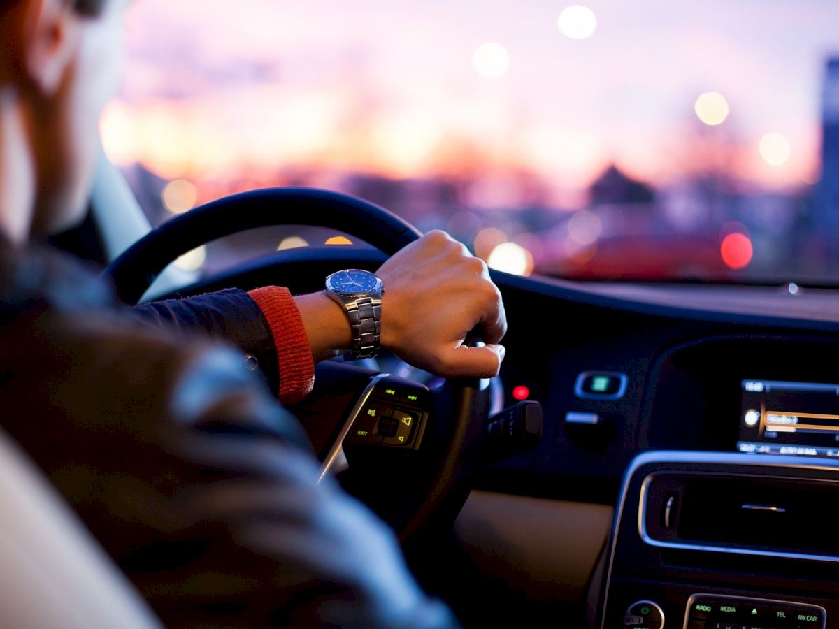 Person driving a car during sunset, holding the steering wheel with their left hand, and dashboard lights visible.