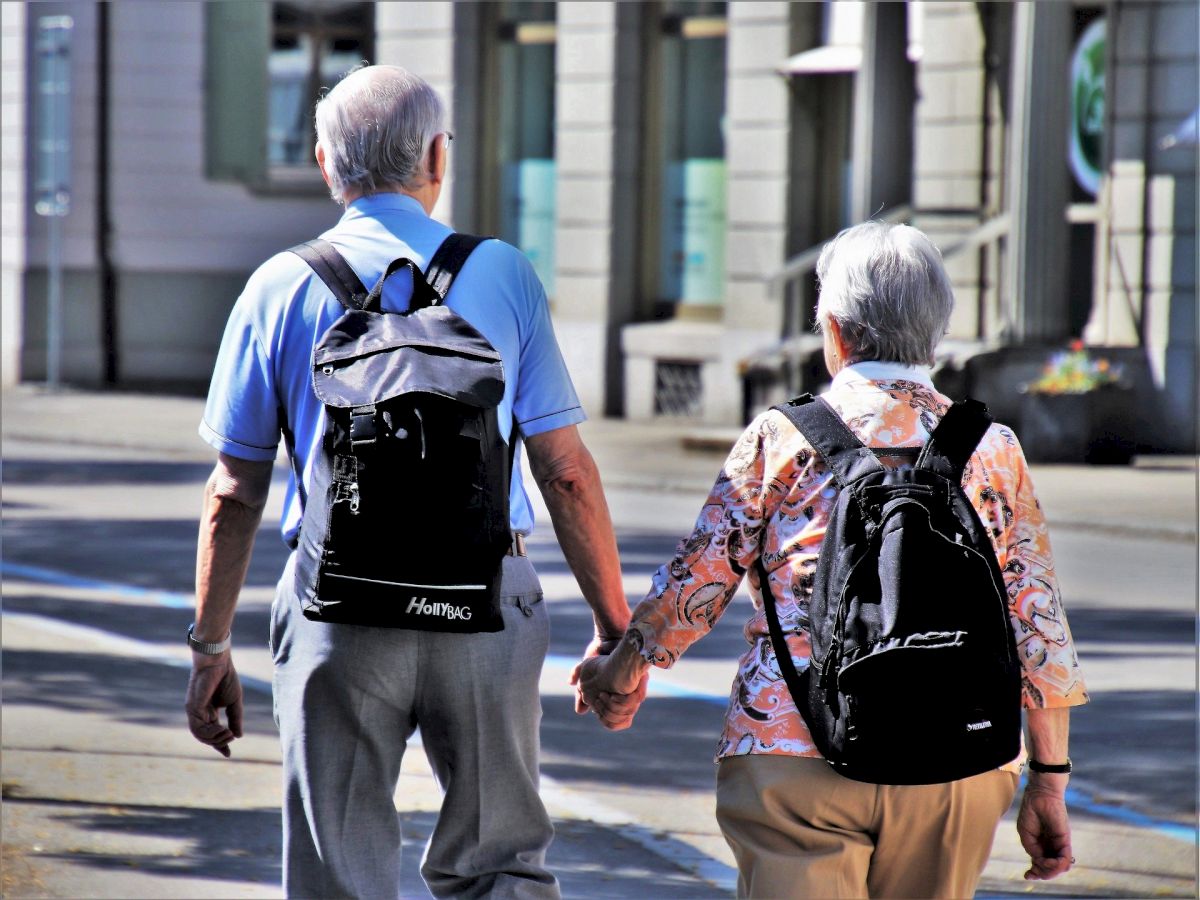 Two elderly people are walking hand-in-hand down the street, each carrying a backpack.