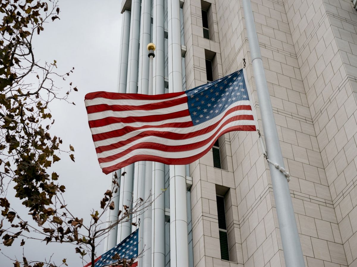 An American flag waves in front of a tall building, beside a tree with autumn leaves, on a cloudy day.