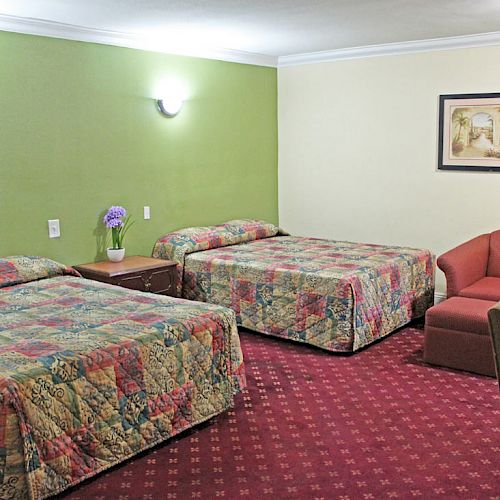 A hotel room with two beds, a red chair, green walls, and colorful bedspreads, also includes a picture on the wall and bedside tables ending the sentence.