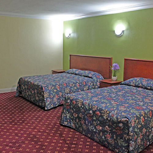 A hotel room with two double beds with floral bedspreads, a nightstand with a flower vase, and wall-mounted lights is shown.