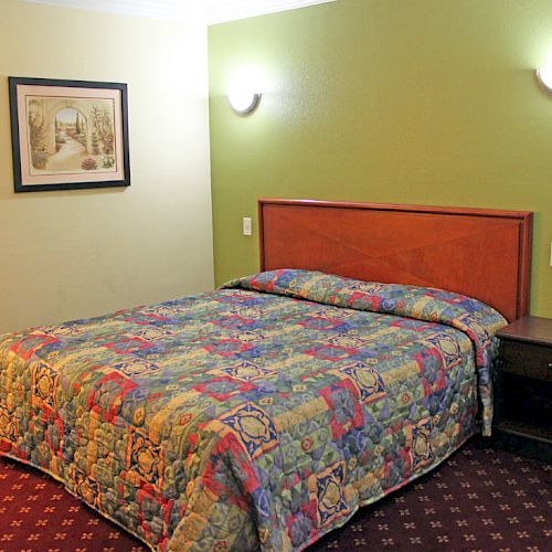 A hotel room with a green wall, queen bed with a colorful quilt, a bedside table, wall lamps, a picture frame, and an open door to a bathroom.