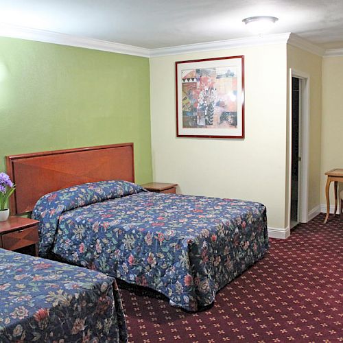 A hotel room includes two beds with floral bedspreads, a painting, a nightstand, a lamp, and a desk with a chair.