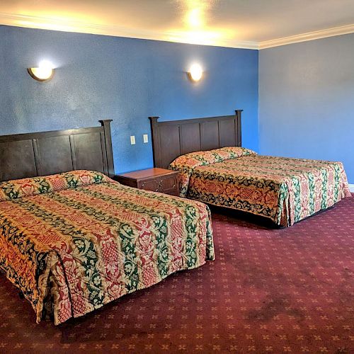 A hotel room with two double beds covered in patterned bedspreads, a nightstand between them, blue walls, and a red carpet.