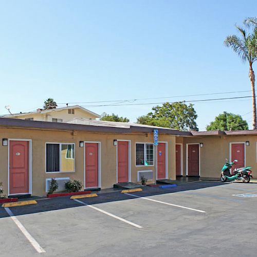 A small motel with pink doors, a few parking spaces, a parked motorcycle, palm trees, and a vending machine on the right end of the building.