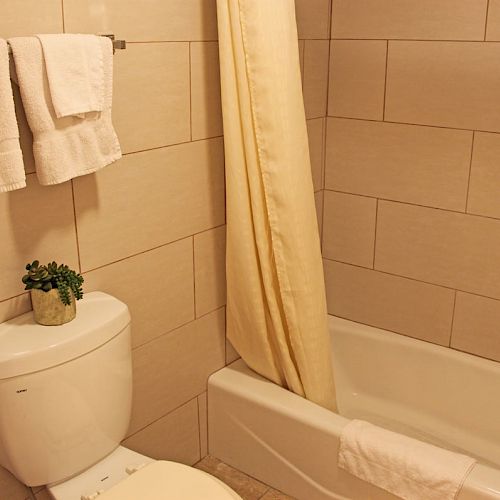 The image shows a clean bathroom with beige tiles, featuring a toilet with a plant on top, a bathtub with a shower curtain, and neatly hung towels.