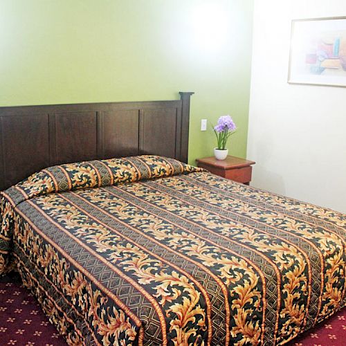 The image shows a bedroom with a large bed that has a patterned bedspread, a green accent wall, a side table with a vase of flowers, and a framed picture.