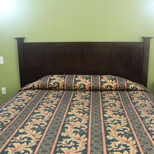 A neatly made bed with a patterned cover, wooden headboard, and a bedside table with a potted plant, set against a green wall.