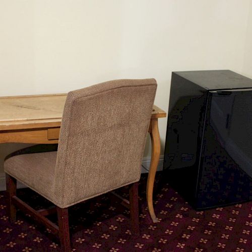 A small workspace with a wooden desk, a cushioned chair, and a mini-fridge on a maroon patterned carpet, all against a beige wall.