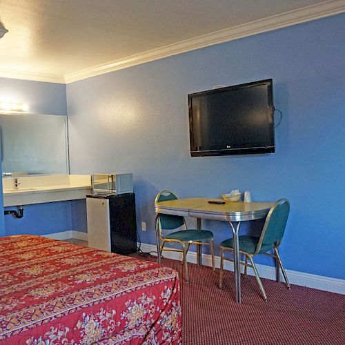 A motel room with blue walls, a red floral bedspread, a wall-mounted TV, a small table with two chairs, and a vanity area with a sink and mirror.