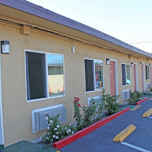 The image shows the exterior of a motel with beige walls, pink doors, small garden beds, and parking spaces with yellow wheel stops.