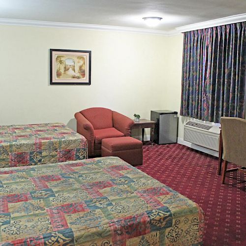 The image shows a hotel room with two beds, an armchair, a desk with a chair, a flat-screen TV, and a small fridge under the desk.