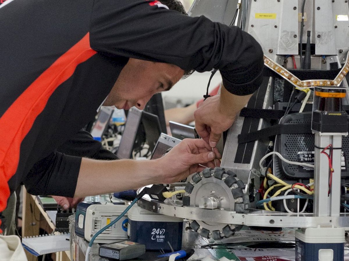 A person is working on a complex machine or robotic equipment, using tools, surrounded by various components and wires.