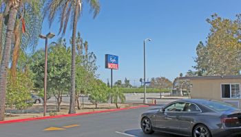 A parked car is in the lot of a motel with the sign visible, surrounded by palm trees and clear skies, ending the sentence.