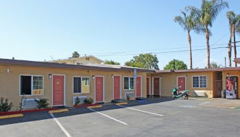 This image shows a small motel with pink doors, a few parking spaces, a motorcycle parked outside, and palm trees in the background.