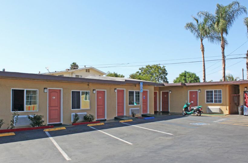 This image shows a small motel with pink doors, a few parking spaces, a motorcycle parked outside, and palm trees in the background.