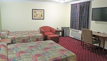 The image shows a hotel room with two double beds, a red chair, a desk with a chair, an air conditioner, a TV, and a window with curtains.