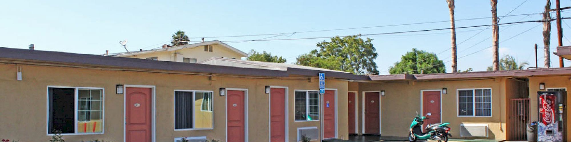 The image shows a small motel with multiple beige rooms, each door painted pink. A scooter is parked outside, with palm trees in the background.