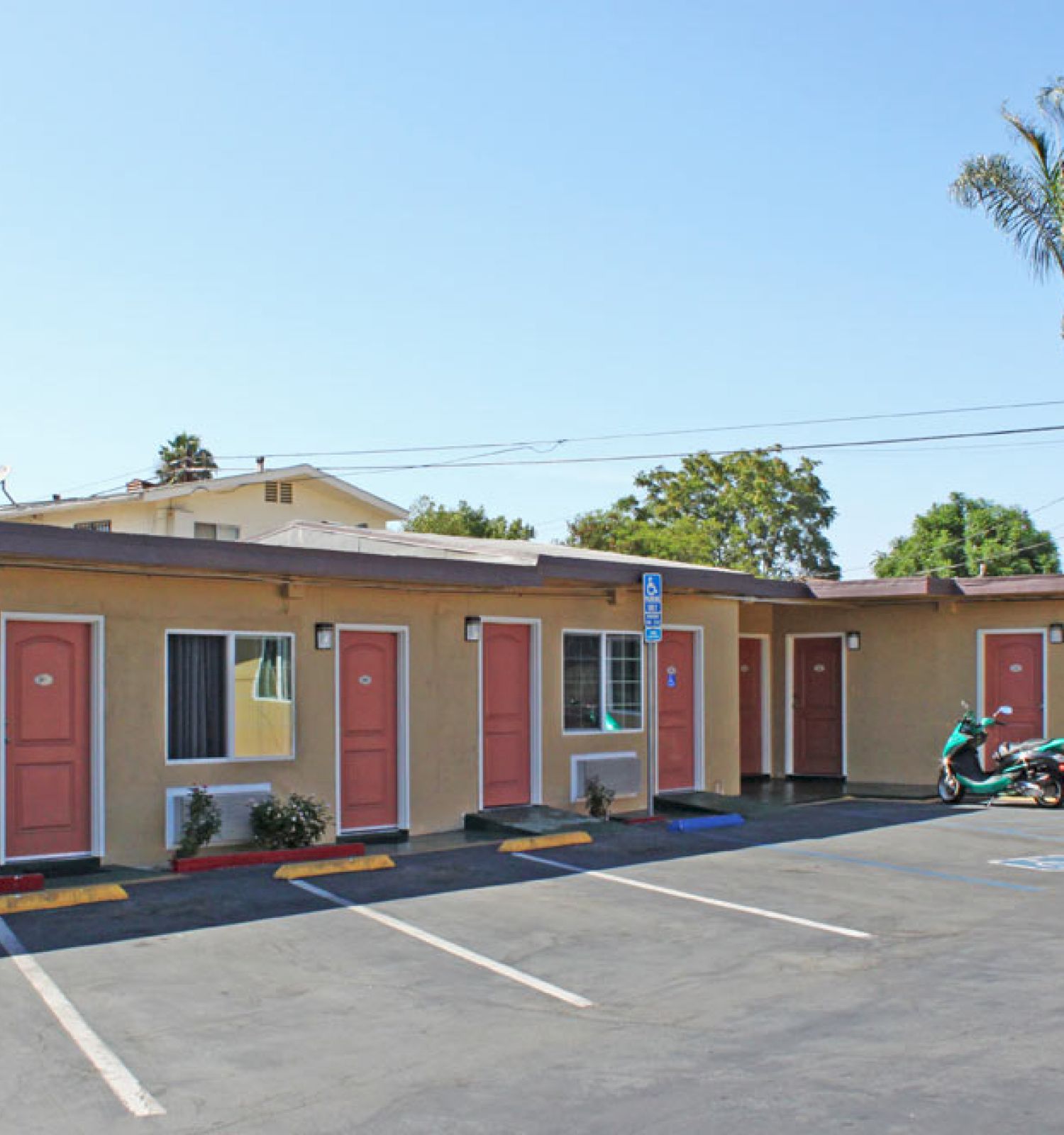 A small motel with a parking lot, pink doors, palm trees in the background, a parked motorbike, and a vending machine to the right.