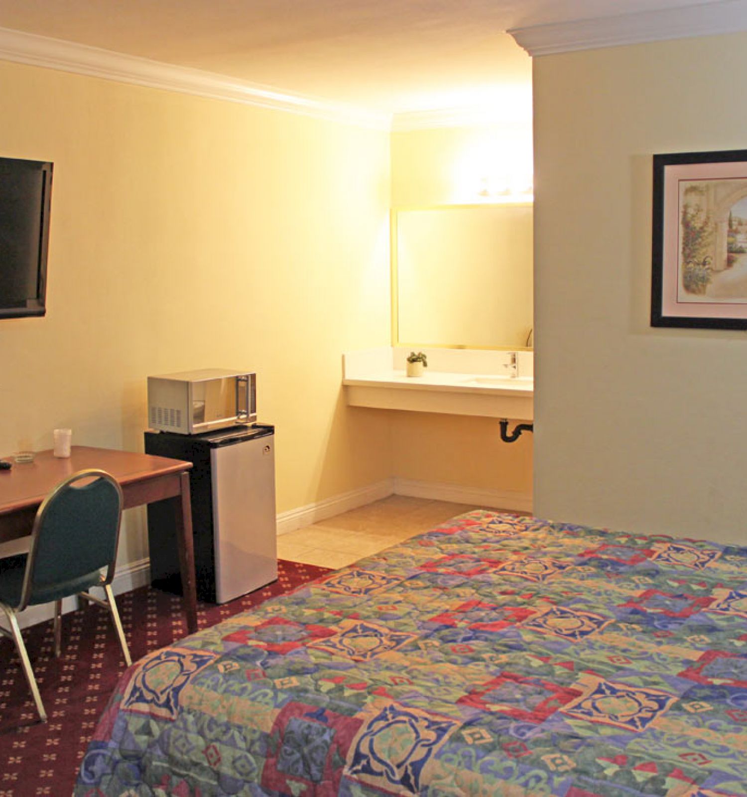 A hotel room with a bed, desk, chair, TV, fridge, and a sink area, decorated with a painting and flowers on the desk, is shown.