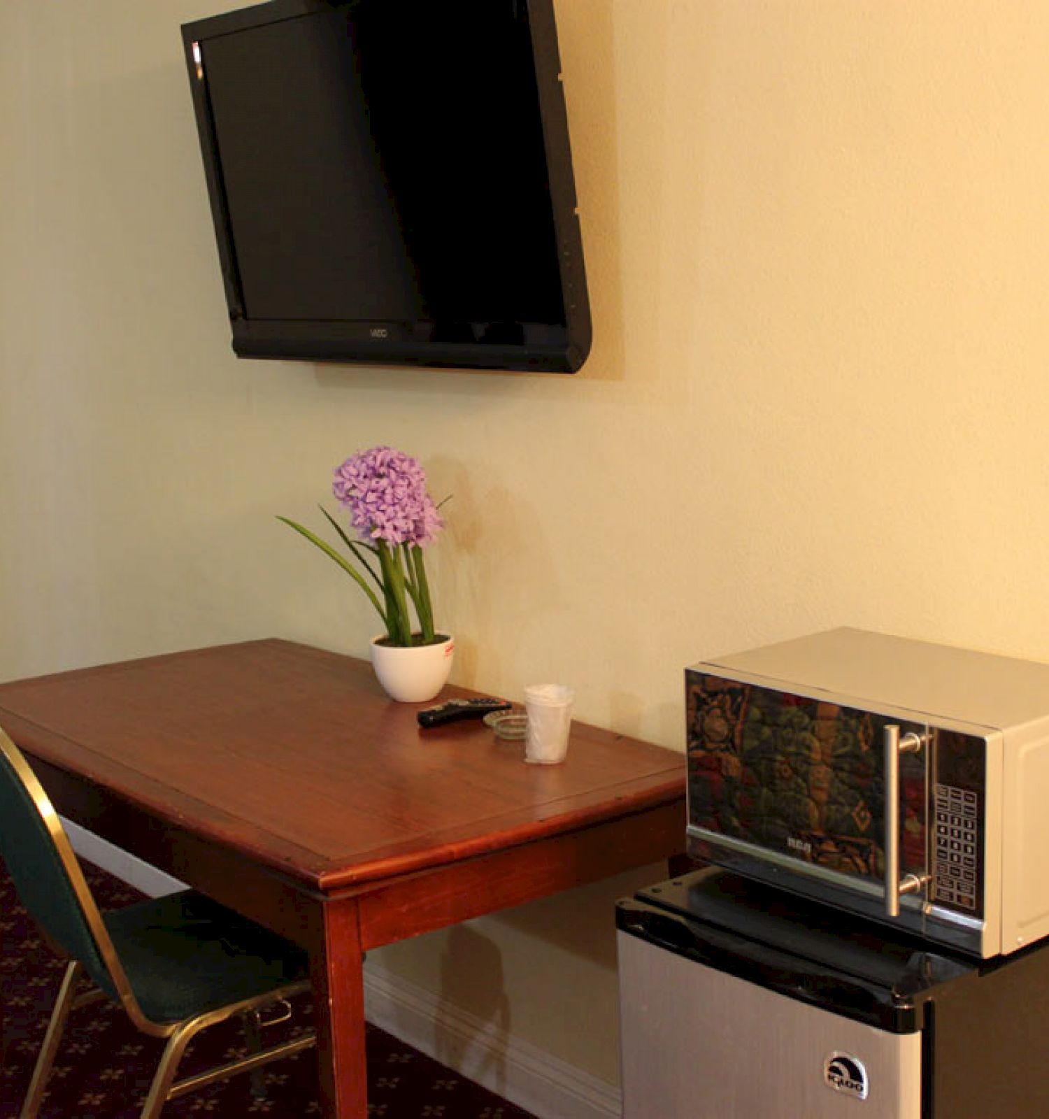 The image shows a hotel room with a wall-mounted TV, a wooden table with a potted plant, a chair, a microwave, and a mini refrigerator.