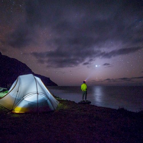 A person in a yellow jacket stands near a lit tent by a lake at night, gazing at a cloudy, star-lit sky with mountains in the background.