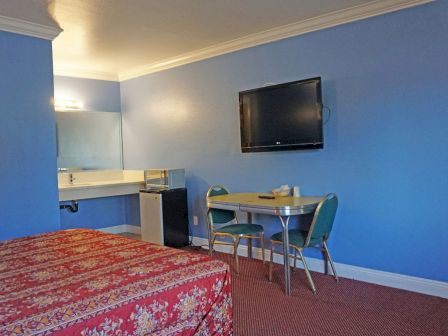 A room with a red floral bed, blue walls, TV, table with chairs, and a small sink area.