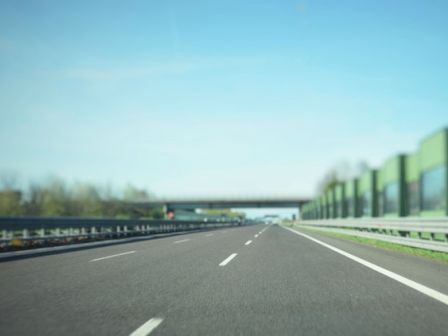 The image depicts an empty highway with multiple lanes, clear skies, trees on the left, and noise barriers on the right, leading to an overpass.