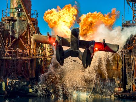 A dramatic scene depicts a seaplane taking off from water with large explosions in the background among industrial structures, creating a thrilling atmosphere.