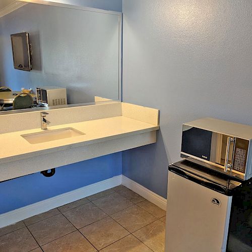 A small kitchenette with a microwave and mini-fridge next to a long counter with a sink and large mirror on a blue wall.